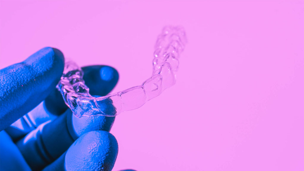 An invisalign dentist hand in a blue glove holds aligners for aligning teeth on a pink background.