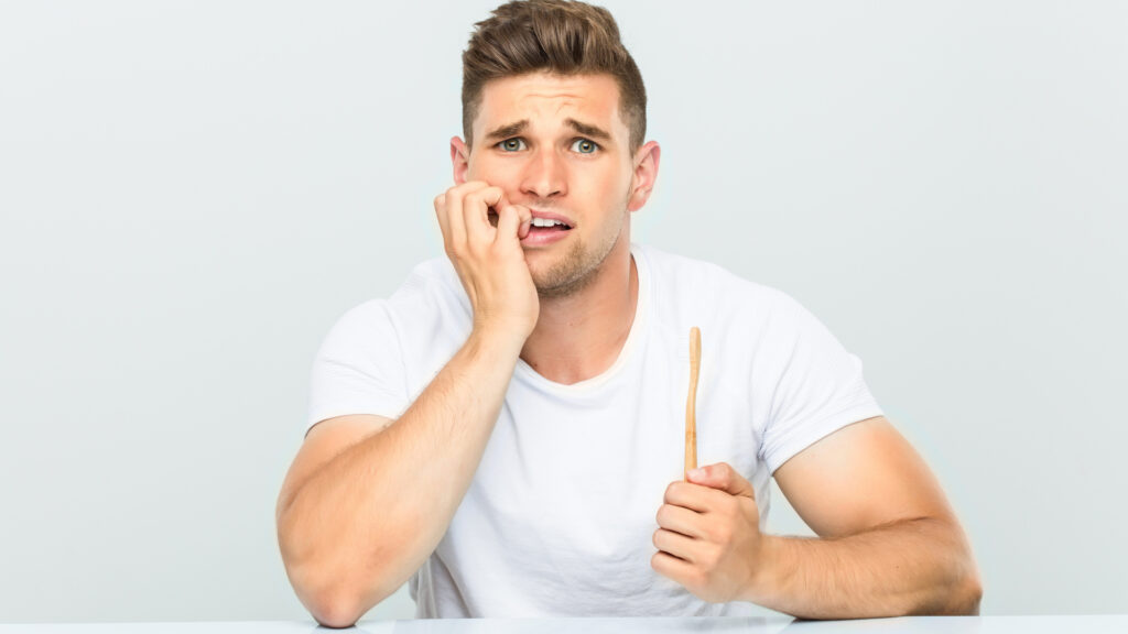 Man biting nails and holding toothbrush as he listens to 5 tips for overcoming dental anxiety