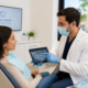 A dentist wearing a mask and gloves shows a dental X-ray on a tablet to a female patient sitting in a dental chair.